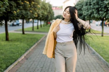 Uzun siyah saçlı, uzun siyah pantolonlu, beyaz gömlekli, sarı ceketli, şehirli, genç, melez Asyalı kadın güneşli bir günde yeşil ağaçlarla şehir sokağında yürüyor.