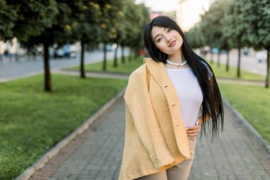 Uzun siyah saçlı, sarı renkli ceketli, yeşil ağaçlarla dolu güzel şehir sokağının arka planında kameraya gülümseyen sevimli Asyalı genç bir kızın portresi.