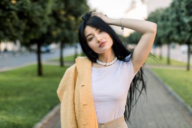 Beyaz tişörtlü, sarı ceketli, mutlu, romantik Asyalı bir kadının portresi. Arka planda yeşil ağaçlarla güzel şehir caddesinde yürüyor.