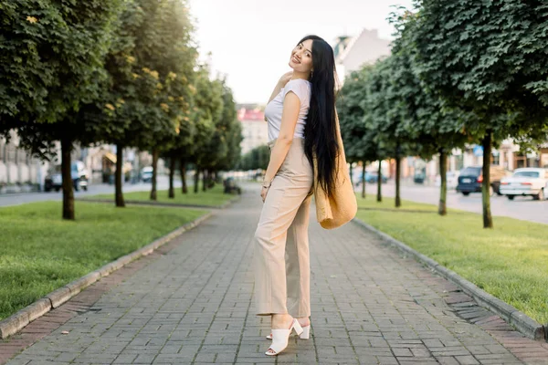 Retrato completo de mulher bonita e elegante jovem com longos cabelos pretos vestindo roupas bege amarelas, fica na estrada no beco da rua na cidade. Retrato da cidade ao ar livre da menina asiática — Fotografia de Stock