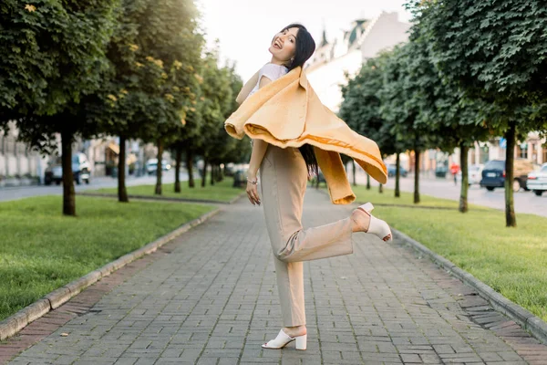 Giovane bella elegante bruna alla moda ragazza caucasica in pantaloni beige e giacca gialla, divertirsi passeggiando all'aperto in città, in posa per la fotocamera con il sorriso al vicolo con alberi verdi — Foto Stock