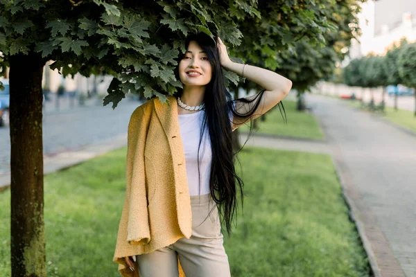 Портрет гарної азіатської дівчини з довгим темним волоссям на вулиці на тлі алеї міського парку з зеленими деревами. Портрет вродливої жінки у модному жовтому світлі, що виходить надвір. — стокове фото