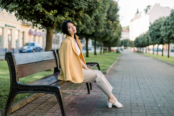 Bella giovane donna d'affari asiatica, indossa elegante giacca gialla casual elegante e pantaloni beige, seduto su una panchina in un vicolo della città tra alberi verdi e godendo l'inizio della giornata di sole — Foto Stock