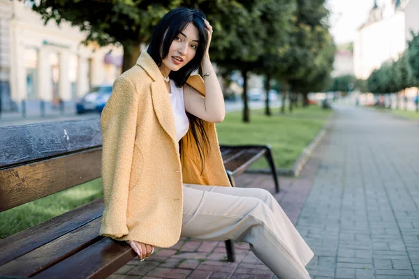Молода жінка азіатська модель з довгим прямим чорним волоссям, одягнена в бежеві штани і жовтий піджак, сидячи на дерев'яній лавці в міському парку алеї, посміхаючись в камеру в сонячний день. — стокове фото