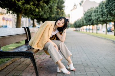 Güzel esmer, melez, seksi kız, modaya uygun sarı ceket ve bej pantolon giyiyor, arka planda yeşil ağaçlarla şehir merkezinde bankta otururken kameraya poz veriyor.