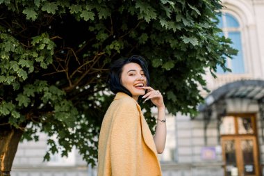 Sarı ceketli, kameraya poz veren ve şehir yürüyüşünün tadını çıkaran güzel melez bir kızın açık hava portresi. Zarif Asyalı kadın, arka planda ağaç ve bina ile bankta oturuyor.