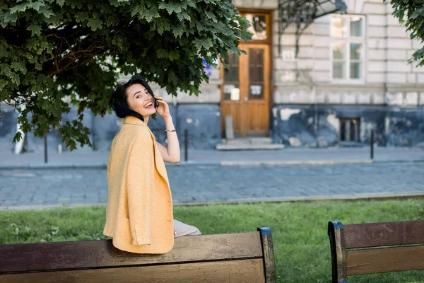 Сміючись елегантно азіатською жінкою в жовтій куртці, охолоджуючи і отримуючи задоволення під час своєї ранкової прогулянки по місту. На вулиці фото прекрасної бізнес-леді з брюнеткою сидить на лавці.. — стокове фото