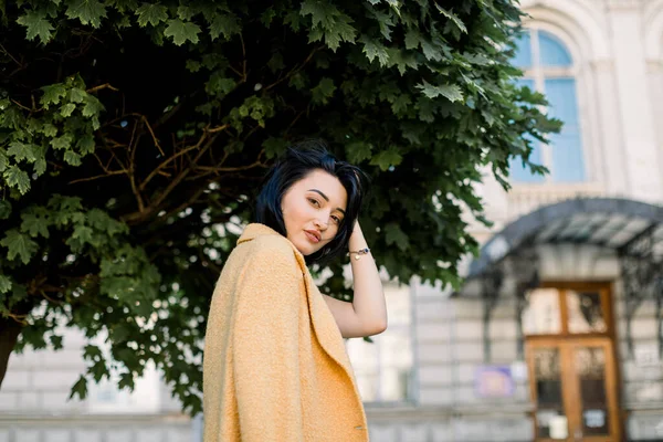 Portrait en plein air de jolie jeune fille blanche métissée en blazer jaune, posant devant la caméra et profitant de sa promenade en ville. Élégante femme asiatique assise sur un banc avec bâtiment de la ville et arbre sur le fond — Photo