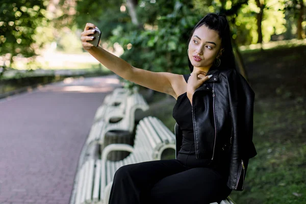 Retrato ao ar livre de menina asiática com estilo de cabelo de rabo de cavalo, vestindo terno preto na moda e jaqueta de couro, sorrindo para a câmera enquanto toma selfie, sentado no banco no belo parque da cidade de verão — Fotografia de Stock