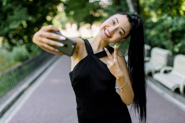 Pretty Asian funny laughing girl with beautiful nude make up and ponytail hair style, showing V sign while taking selfie in park outdoors. Emotions, people, races, portraits concept