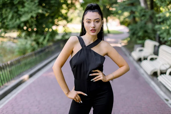 Emotional outdoor portrait of charming young Asian Japanese girl with black ponytail hair, wearing trendy black overalls, posing to camera, enjoying summer walk in the park. Front view — Stock Photo, Image