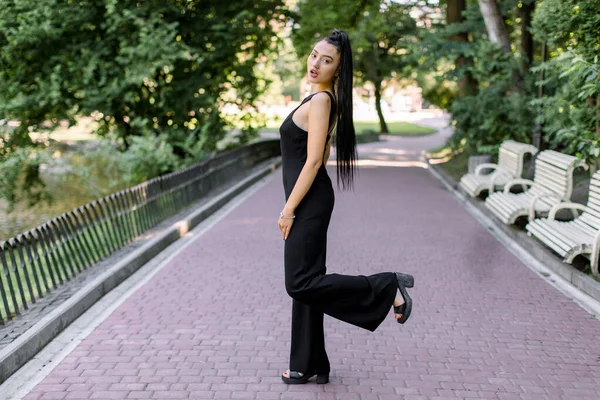 Retrato al aire libre de una hermosa mujer asiática sexy de raza mixta vestida con monos negros, posando en el callejón del parque durante el paseo al atardecer, con una pierna levantada. Linda chica morena en ropa elegante en el parque —  Fotos de Stock
