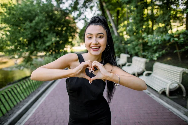 Apaixonado pelo mundo. Retrato de close-up da menina asiática chinesa sorridente moderna mostrando as mãos em forma de coração para a câmera, enquanto estava fora no verão verde árvores cidade parque fundo — Fotografia de Stock