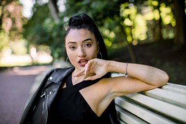 Parktaki bankta oturan, siyah elbise ve deri ceketli, uzun at kuyruklu, güzel seksi Çinli kadının yarım boy yatay portresi. İnsanlar, duygu konsepti