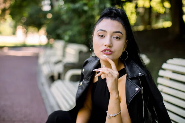 Retrato horizontal de meio comprimento de bela sensual sonhadora jovem mulher asiática, vestindo roupa preta na moda, com cabelo longo rabo de cavalo escuro, sentado em um banco em um parque. Pessoas, conceito de emoções — Fotografia de Stock