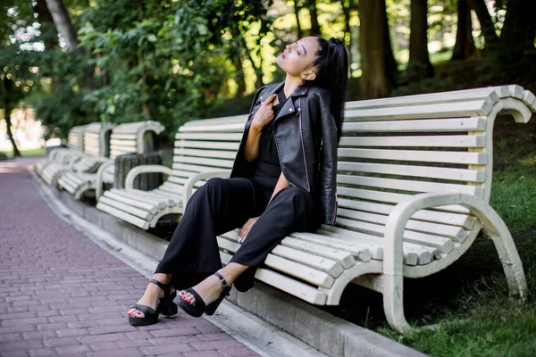 Lifestyle portrait en plein air de heureuse jeune belle femme asiatique mixte, portant un pantalon noir et une veste en cuir, assis sur un banc dans un beau parc vert. Concept de personnes, de marche et de loisirs — Photo