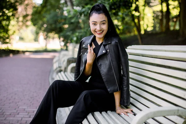 Retrato de jovem mulher asiática muito rindo na moda calças pretas e jaqueta de couro, sentado no banco verde do parque da cidade. Estilo de vida, lazer e conceito de pessoas. — Fotografia de Stock