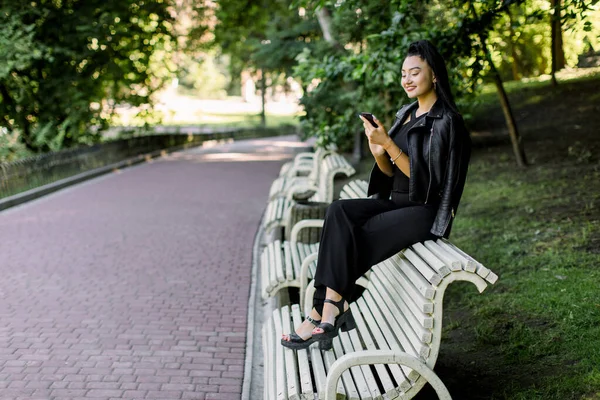 Дуже радісна молода азійка, одягнена в модний чорний одяг, сидить на лавці в міському парку, тримає мобільний телефон, читаючи новини, соціальні мережі або набираючи повідомлення. — стокове фото