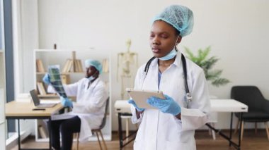 Tıbbi ekip üyeleri görev başında. Cerrahi başlık, eldiven, steteskop takan kadın doktor, tablet üzerinde inceleme notları. Bilgisayarda arka plandaki meslektaşım. Profesyonel sağlık hizmeti kavramı.