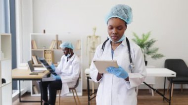 Koruyucu ekipman giyen Afrikalı kadın doktor elinde tablet cihazıyla klinikte dikiliyor. Arka plan hemşiresi masasında beyaz önlüklü çalışıyor..