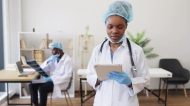 Tıbbi üniformalı genç bayan doktor ve eldiven tutan cihaz, arkada çalışan iş arkadaşı. Modern ofiste sağlık ve profesyonelliği vurgulayan tıbbi personel.