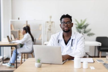 Tıp bürosunda laptopta çalışan beyaz önlüklü profesyonel siyahi erkek doktor. Çalışma alanı profesyonel araçlar, yazma materyalleri ve tıbbi kaynaklar içerir.