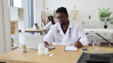 20 'li yaşlarda doktor olarak çalışan kendinden emin bir Afrikalı adam masasında steteskopla çalışıyor. Modern tıbbi ofis ortamında uzmanlık ve profesyonelliği sağlık ve iletişim üzerine odaklıyor.
