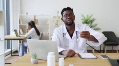 Elinde bir bardak su tutan beyaz önlüklü erkek doktor klinik ortamında sağlık eğitiminin içme suyu faydalarını vurguluyor. Hidrasyon önemini gösterir, izleyicileri sağlığa bağlar