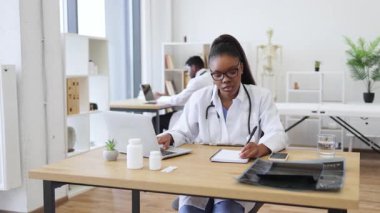 Laptop kullanan ve modern tıp ofisinde reçete yazan kendine güvenen genç bir kadın doktor. Sağlık ekipmanlarının yer aldığı parlak çalışma alanı, arka planda meslektaşla birlikte resmedilen takım çalışması