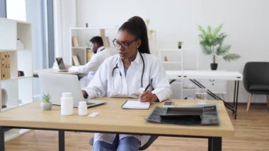 Genç Afrikalı doktor araştırma için dizüstü bilgisayar kullanıyor, not defterine notlar yazıyor. İşyerindeki profesyonellik tıbbı ve teknolojisini yansıtıyor. Arkaplan özellikleri çağdaş iç mimari