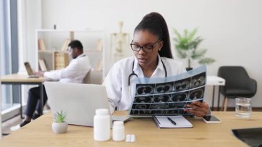 Genç Afrikalı Amerikalı kadın doktor, erkek meslektaşının yanında modern tıp ofisinde röntgen taramasını inceliyor. Profesyonel sağlık, teşhis, işbirliği ortamı sunar.