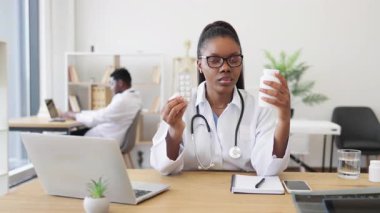 Beyaz önlüklü, steteskoplu, su toplayan siyah bir kadın doktor ve bir bardak su. Profesyonel tıbbi ofis ortamında oturuyorlar..