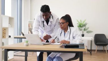 Beyaz önlüklü iki tıp uzmanı modern ofiste dizüstü bilgisayarla hasta tedavi stratejisini tartışıp planlıyorlar. Takım çalışması, araştırma, hasta bakımı kavramlarına odaklan.