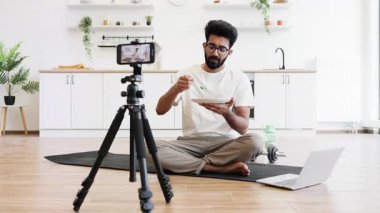 Sakallı genç bir adam, mutfak ortamında sağlıklı beslenmeyle ilgili bir video bloğu kaydediyor. Salata, tripod ve dizüstü bilgisayarla yere oturmuş, sosyal medya izleyicileri için ilgi çekici içerikler yaratıyor..