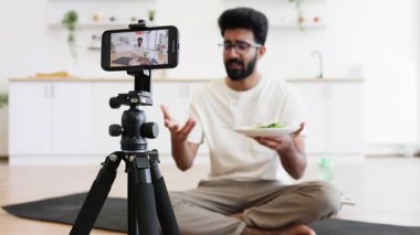 Genç sakallı adam modern apartmanda oturmuş sağlıklı beslenmeyle ilgili video kaydediyor..