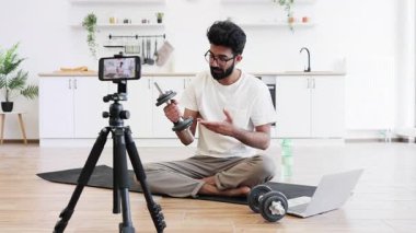 Adam evde yoga minderinde oturmuş halterli ve tripod kameralı bir blog kullanıyor. Konsept, evde kaydedilen fitness videosunu destekleyen sağlık, fitness ve modern yaşam tarzını ön plana çıkarıyor