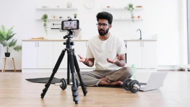 Adam mutfakta video kaydı sırasında dambıllarla egzersiz yapıyor, spor salonunu ve sağlığını gösteriyor. Bilgisayarın ve telefonun yanındaki mindere oturmuş, ev sağlığı motivasyonunu arttırıyor..