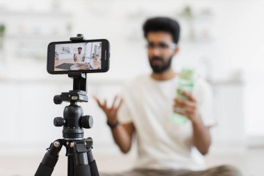 Sakallı, spor yaparken susuzluğun değerini tartışan yetişkin bir adam. Video kurulumu, modern bir mutfak ortamında tripodlu akıllı telefonu içerir..