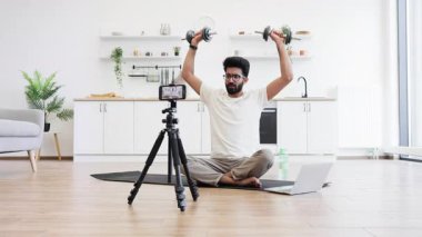 Adam mutfakta video kaydı sırasında dambıllarla egzersiz yapıyor, spor salonunu ve sağlığını gösteriyor. Bilgisayarın ve telefonun yanındaki mindere oturmuş, ev sağlığı motivasyonunu arttırıyor..