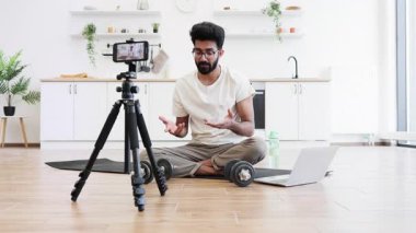 Adam mutfakta video kaydı sırasında dambıllarla egzersiz yapıyor, spor salonunu ve sağlığını gösteriyor. Bilgisayarın ve telefonun yanındaki mindere oturmuş, ev sağlığı motivasyonunu arttırıyor..