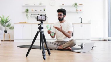 20 'li yaşlarda genç bir adam mutfakta oturmuş egzersiz için suyun önemi hakkında video kaydediyor. Sahne minder, tripod kamera, dambıl içerir, ev ortamında uygunluk içeriğini gösterir.
