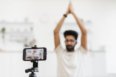 Genç bir adam evde meditasyon dersleri kaydediyor. Akıllı telefon kullanıyor, sağlığını ve farkındalığını vurguluyor. Modern teknolojiyle zihinsel ve fiziksel sağlıkla ilgili içeriği paylaşmanın mükemmel bir örneği