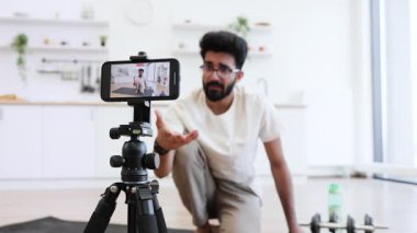 Genç adam kapalı alanda tahta pozu veriyor, modern ekipmanlarla sağlık meraklıları için video dersleri kaydediyor. Görüntü, yumuşak ışık ve özenle döşenmiş mutfağı gösteriyor..