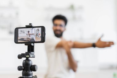 Sakallı yetişkin bir adam spor vlog 'u çekiyor, modern iç mekanlarda kollarını uzatıyor. Akıllı telefon kayıt içeriği üzerine dinamik odak sağlık gösterisi, esneme egzersizleri, spor motivasyonu