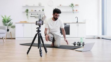 Genç sakallı bir adam evde şınav çekerken yoga minderinde egzersiz yapıyor. Yakınlardaki kamera ve dizüstü bilgisayar sağlık ve sosyal medya içeriğine adanmışlığı gösteriyor.