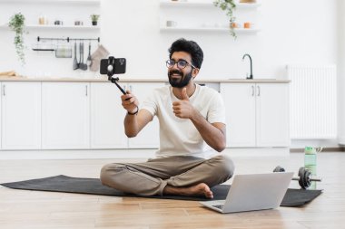 Genç yetişkin erkek tripodda akıllı telefon tutuyor ve evde otururken video kaydediyor.