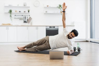 Günlük kıyafetlerle gezen genç sakallı adam evde yoga yapıyor. Blog için sağlık videosu kaydetmek için dizüstü bilgisayar kullanıyor. Spor aletleri ve modern dekor sağlıklı yaşam tarzını ön plana çıkarıyor