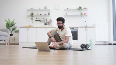 Genç yetişkin bir adam evdeki spor ve spor hakkında video blogu kaydediyor yoga minderinde oturuyor, dizüstü bilgisayarını gösteriyor. Sağlıklı yaşam tarzının içeriğini yakalamak, evde mutfak düzeninde..