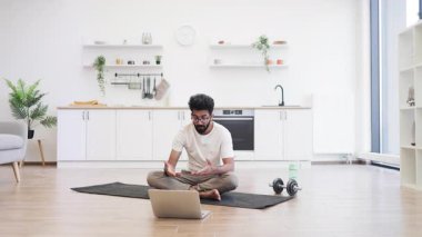 30 'lu yaşlarda genç bir adam, evinde yoga dersi çekerken fitness aktivitelerine katılıyor. Bilgisayarla egzersiz tekniklerini gösteriyor, kişisel refah ve sağlıklı yaşama odaklanıyor.