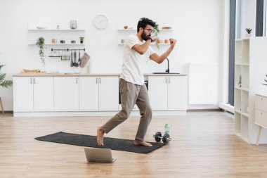 Genç adam boks hareketleri yapıyor. Parlak mutfakta paspaslı yumruklarla. Günlük kıyafetlerle spor yapmak, aktif yaşam tarzını ve sağlığını temiz, modern, kapalı bir ortamda geliştirmek..